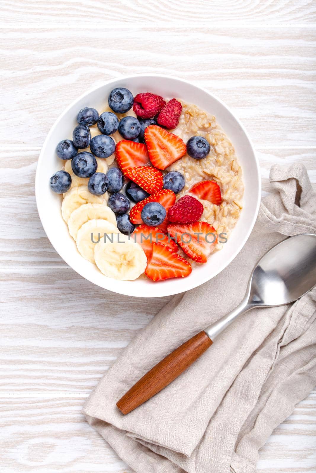 Oatmeal porridge with fruit and berries in bowl with spoon on white wooden background table top view, homemade healthy breakfast cereal with strawberry, banana, blueberry, raspberry. Space for text