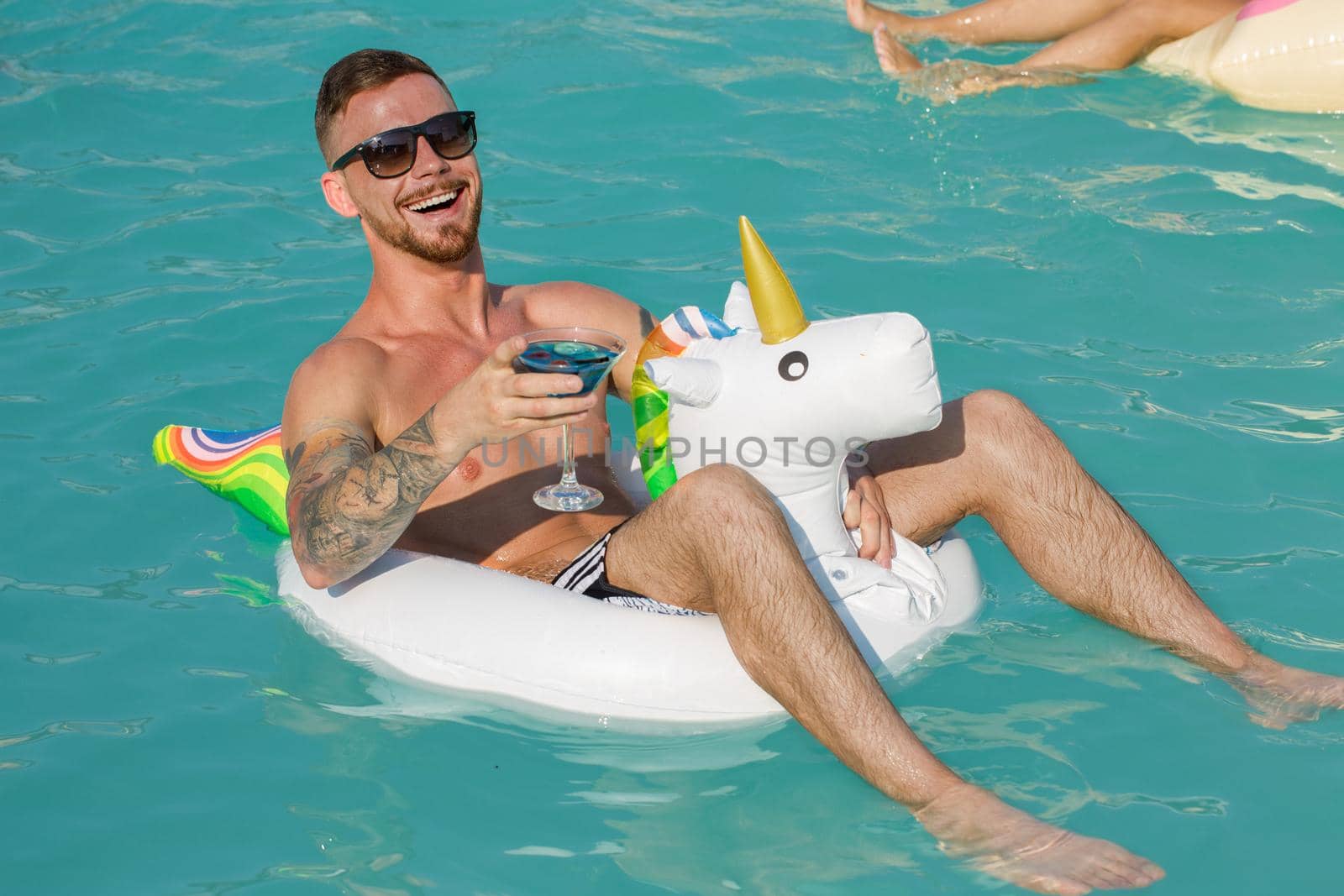 Handsome young tattooed man laughing joyfully swimming on inflatable unicorn at the pool. Cheerful man holding up his drink, enjoying day at the pool, relaxing on inflatable ring. Vacation, travel concept