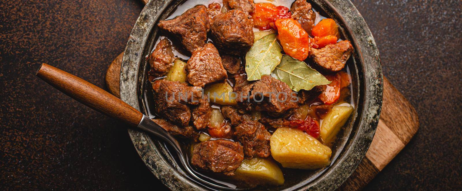 Macro close-up of delicious beef meat stew dish with meat cubes, potatoes, carrot and gravy in rustic metal bowl on brown concrete background top view