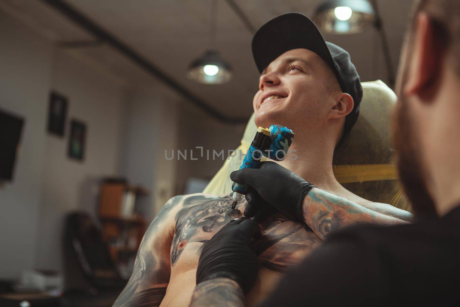 Low angle shot of a young cheerful handsome man smiling joyfully while getting a new tattoo on chest. Attractive man enjoying getting tattooed by professional tattooist