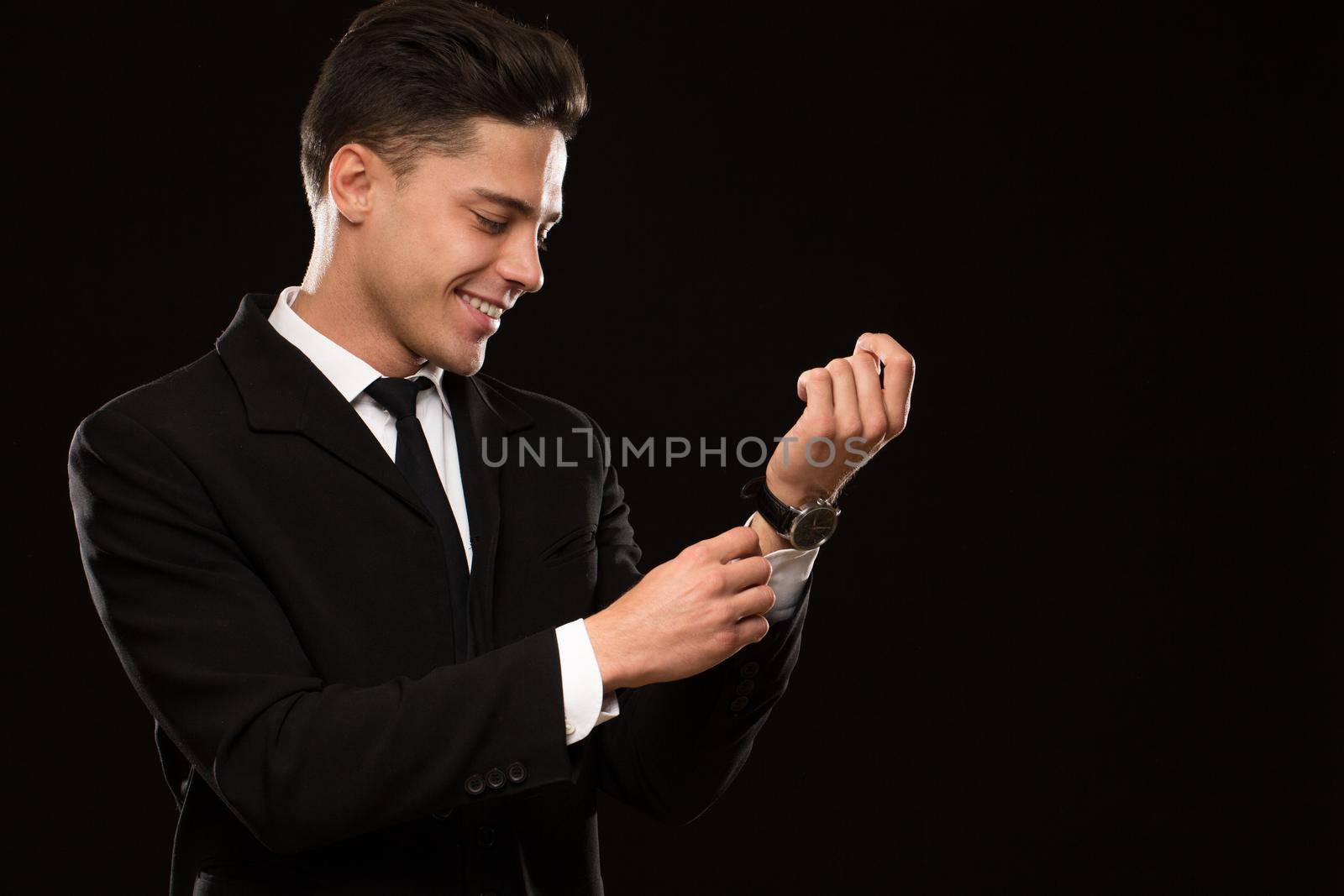 Cheerful handsome young man smiling joyfully adjusting sleeves of his shirt wearing elegant classic black suit and tie on black background copyspace sexy seductive masculinity success