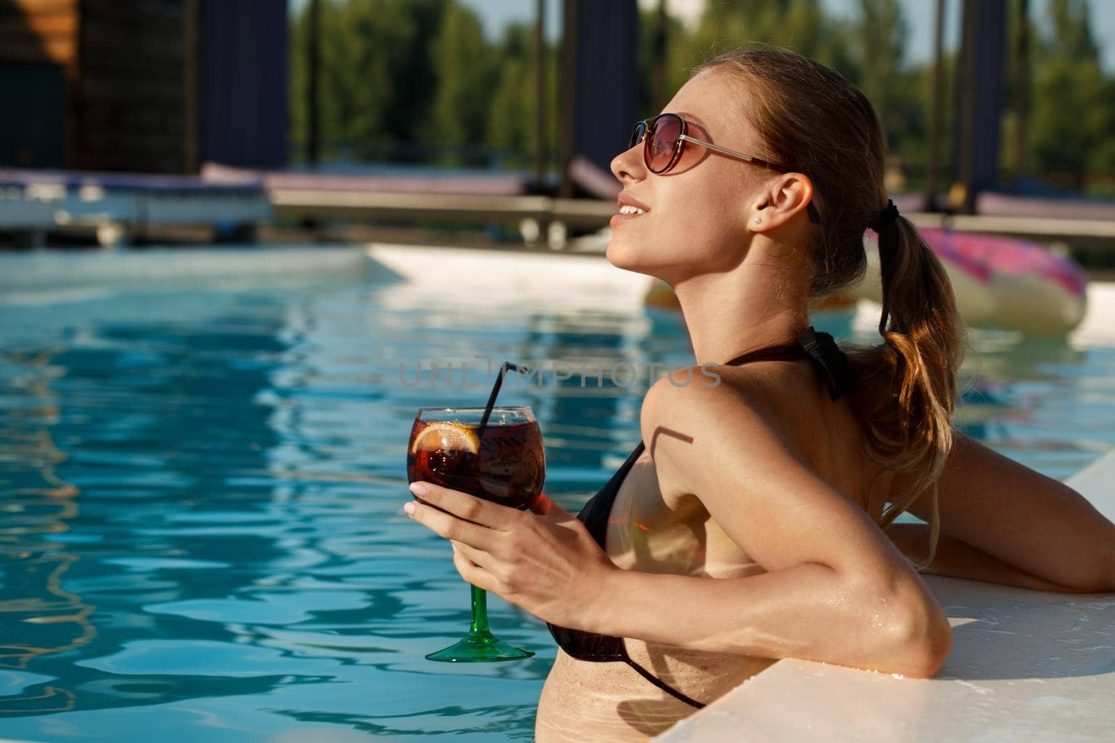 Stunning beautiful young woman enjoyoing sunbathing in the pool with a drink in her hand, copy space. Cheerful attractive woman having cocktail at the poolside. Relation, lifestyle, summertime concept