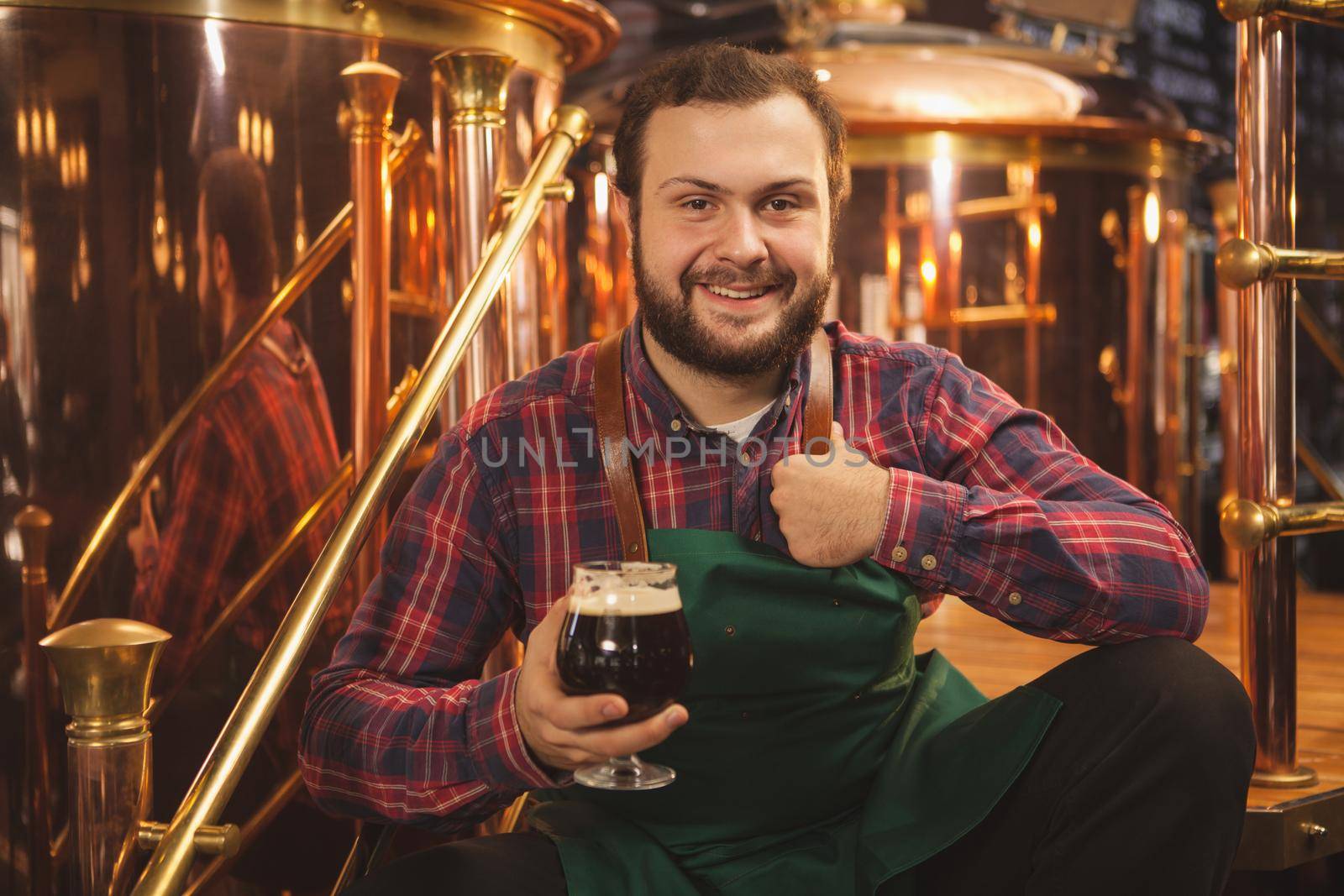 Happy young bearded brewer wearing apron sitting relaxed at his brewery, holding glass of beer showing thumbs up, copy space. Cheerful friendly beermaker enjoying craft beer at his pub