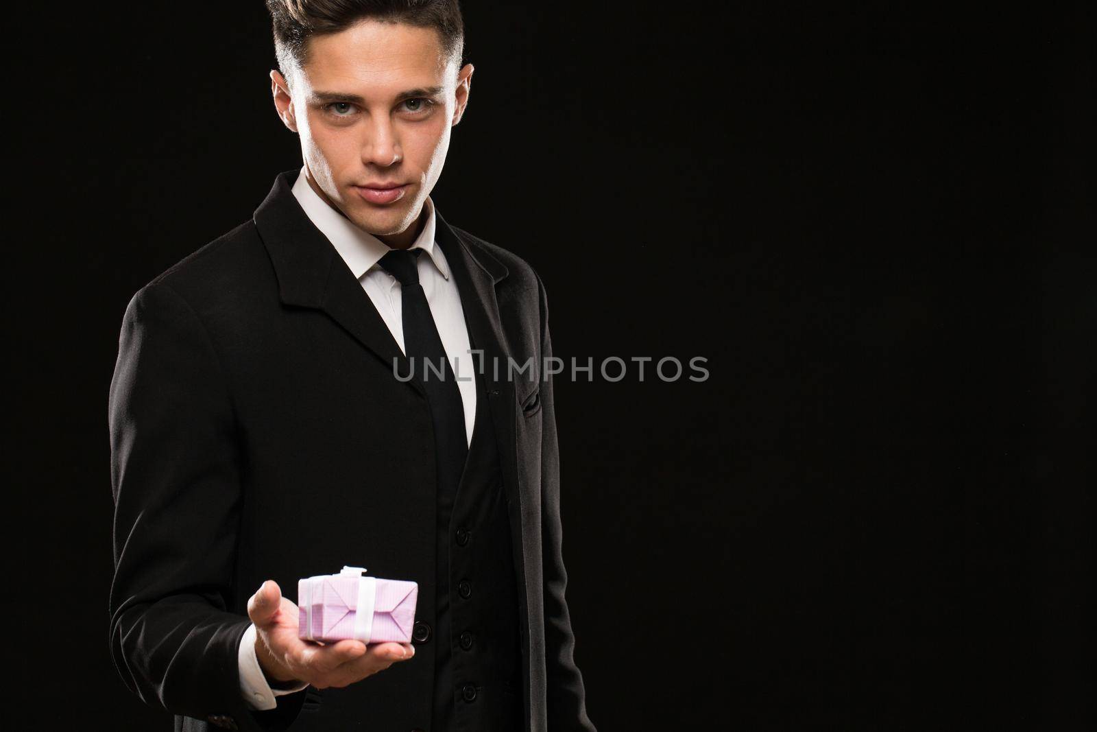 Portrait of a handsome young macho man wearing elegant black suit smiling seductively holding out a small present box to the camera copyspace romance romantic flirt flirtatious anniversary boyfriend