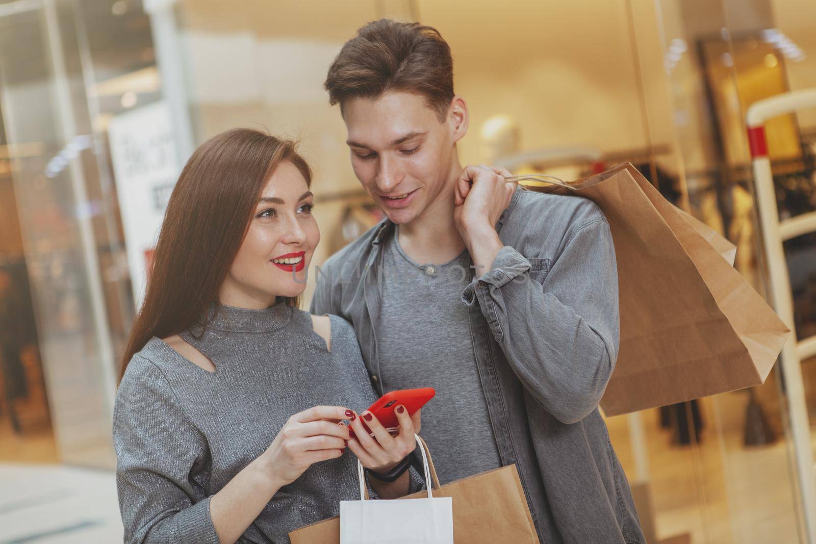 Happy couple using smart phone, while shopping at the mall together. Beautiful woman deciding where to shop, holding her smart phone, browsing online.Boyfriend and girlfriend enjoying seasonal sales