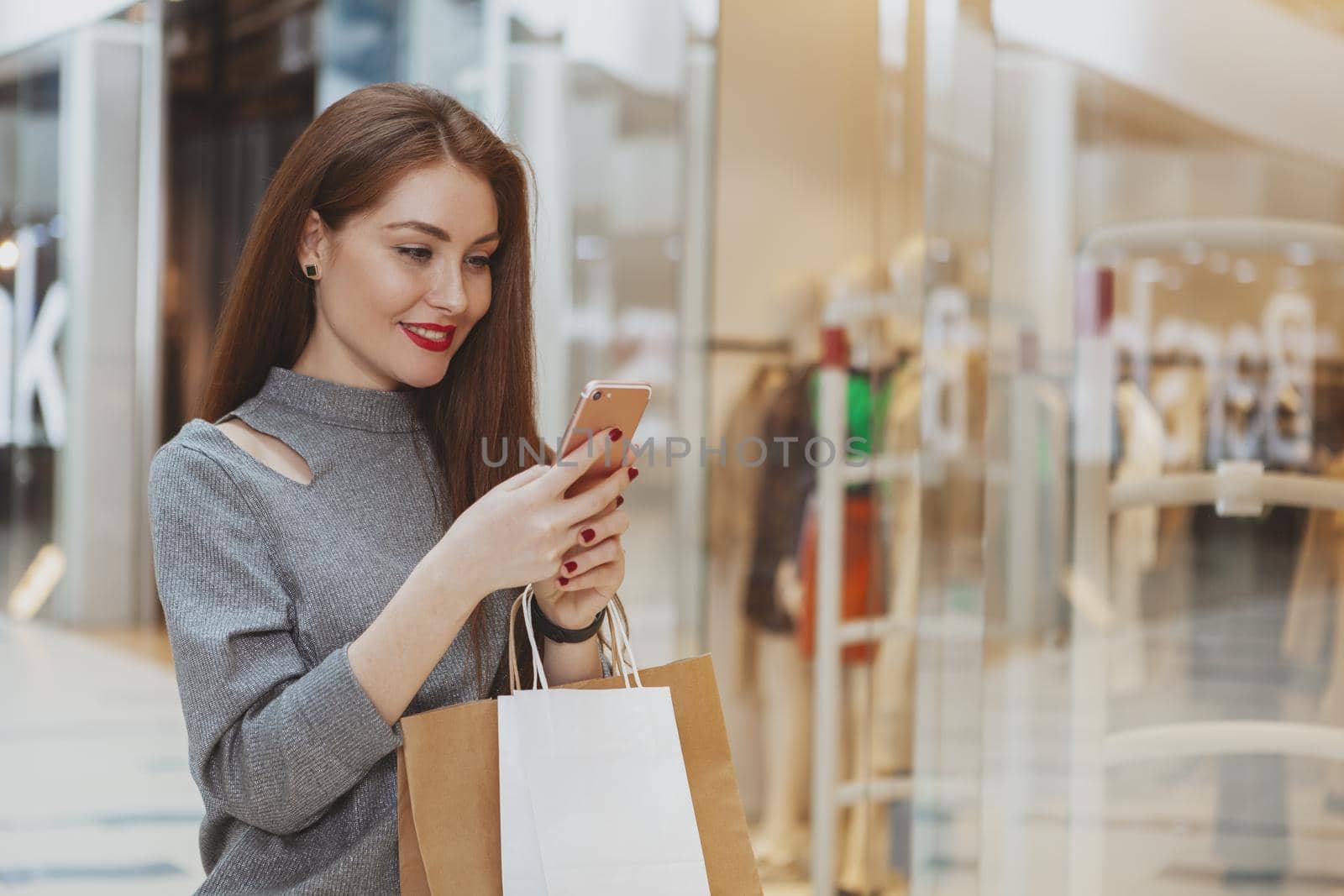 Attractive cheerful female customer using smart phone while shopping at the mall, carrying paper bags with purchase, copy space. Online shopping, application concept