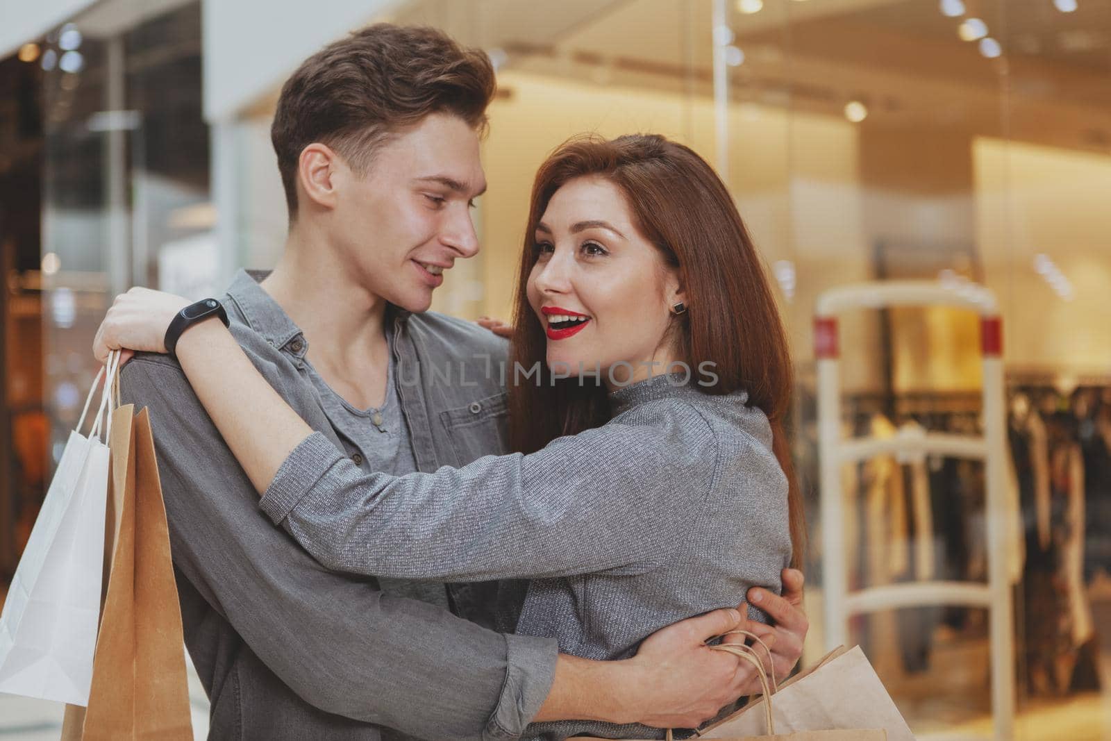 Cheerful loving young man hugging his beautiful girlfriend at the shopping mall. Young woman looking excited, while shopping at the mall with her boyfriend, copy space