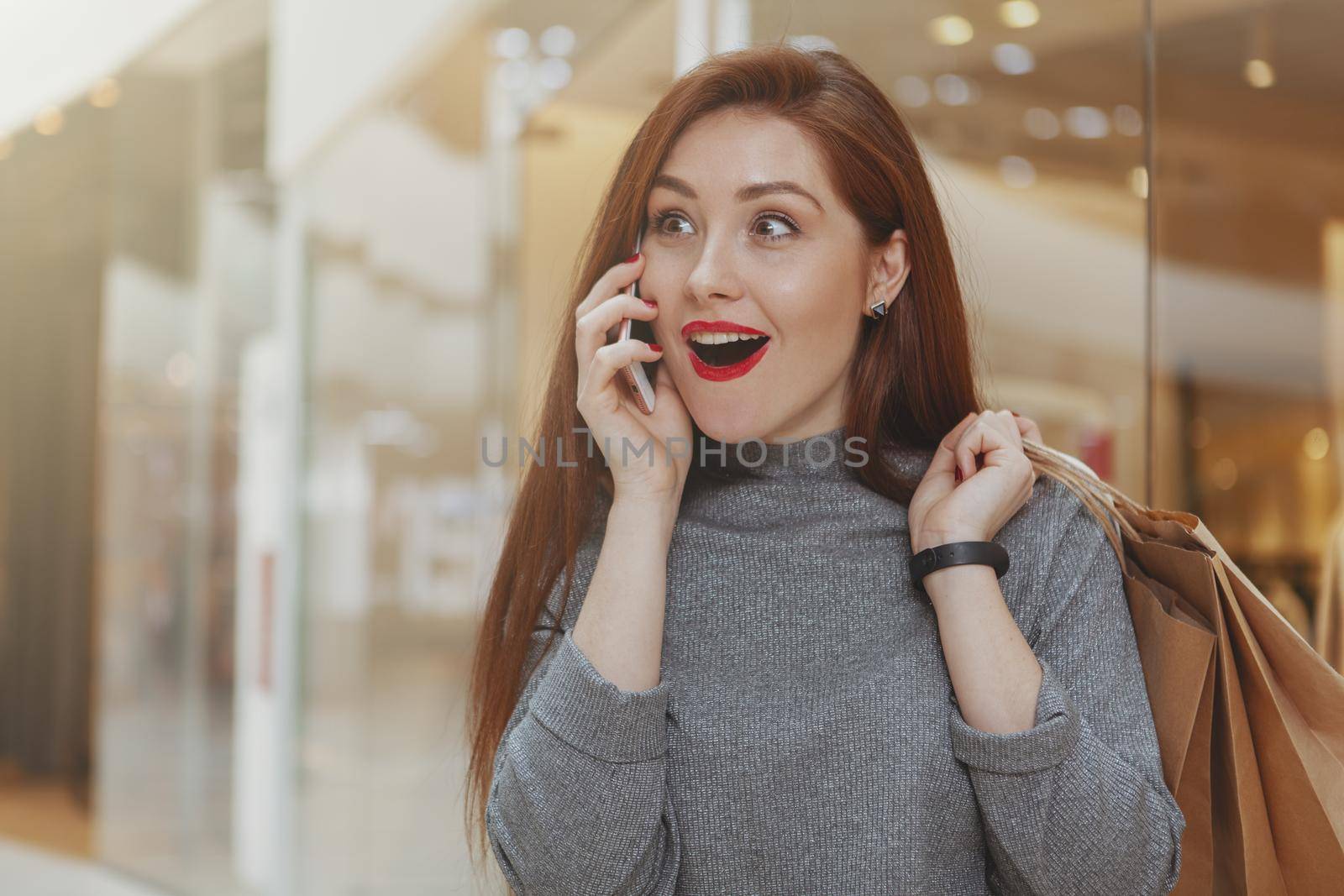 Beautiful young woman looking shocked, talking on her smart phone at the shopping mall, copy space. Gorgeous expressive female customer looking surprised, receiving news call on her mobile