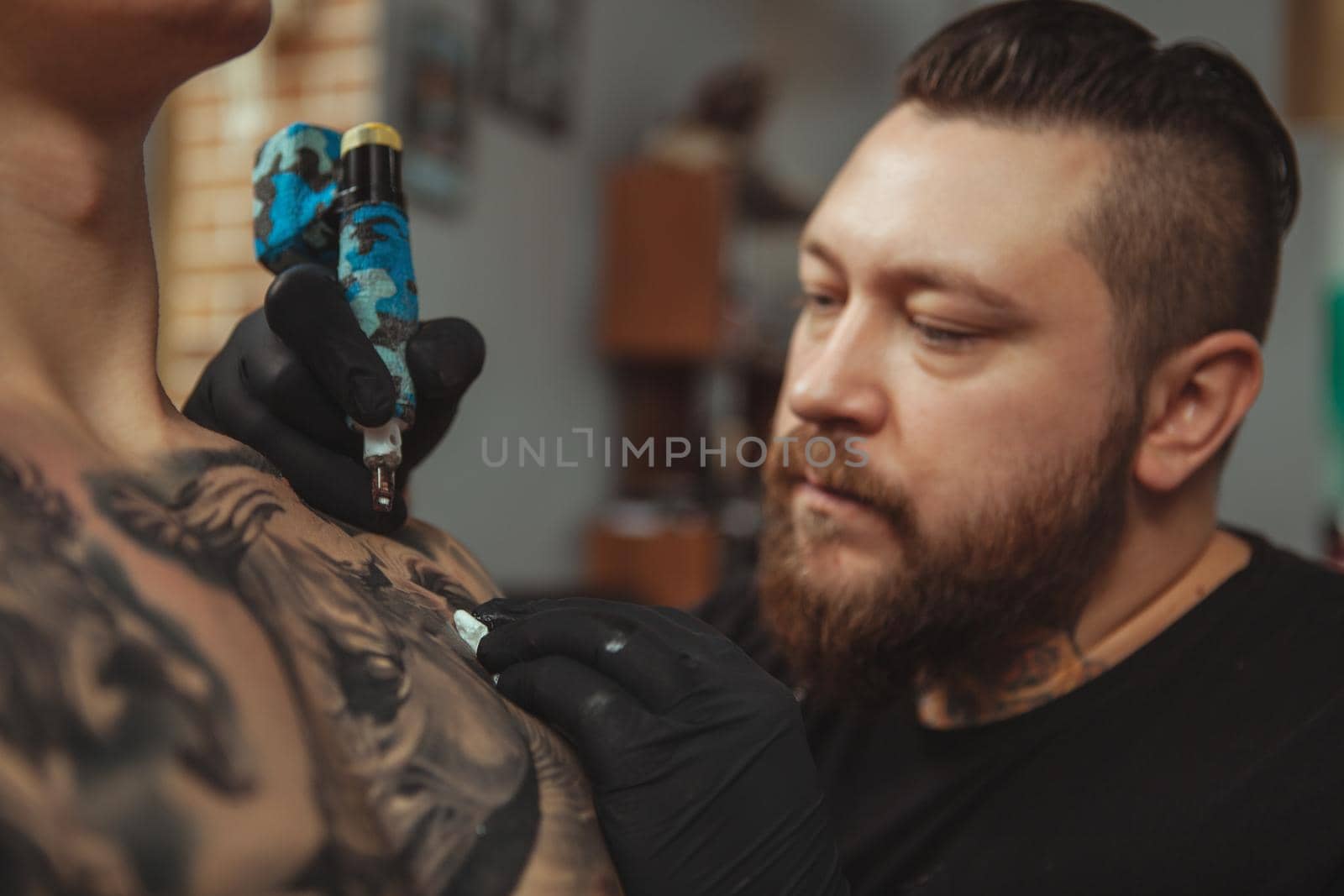 Close up of a bearded professional tattoo artist looking focused, tattooing male client on the chest, copy space. Male tattooist working at his studio