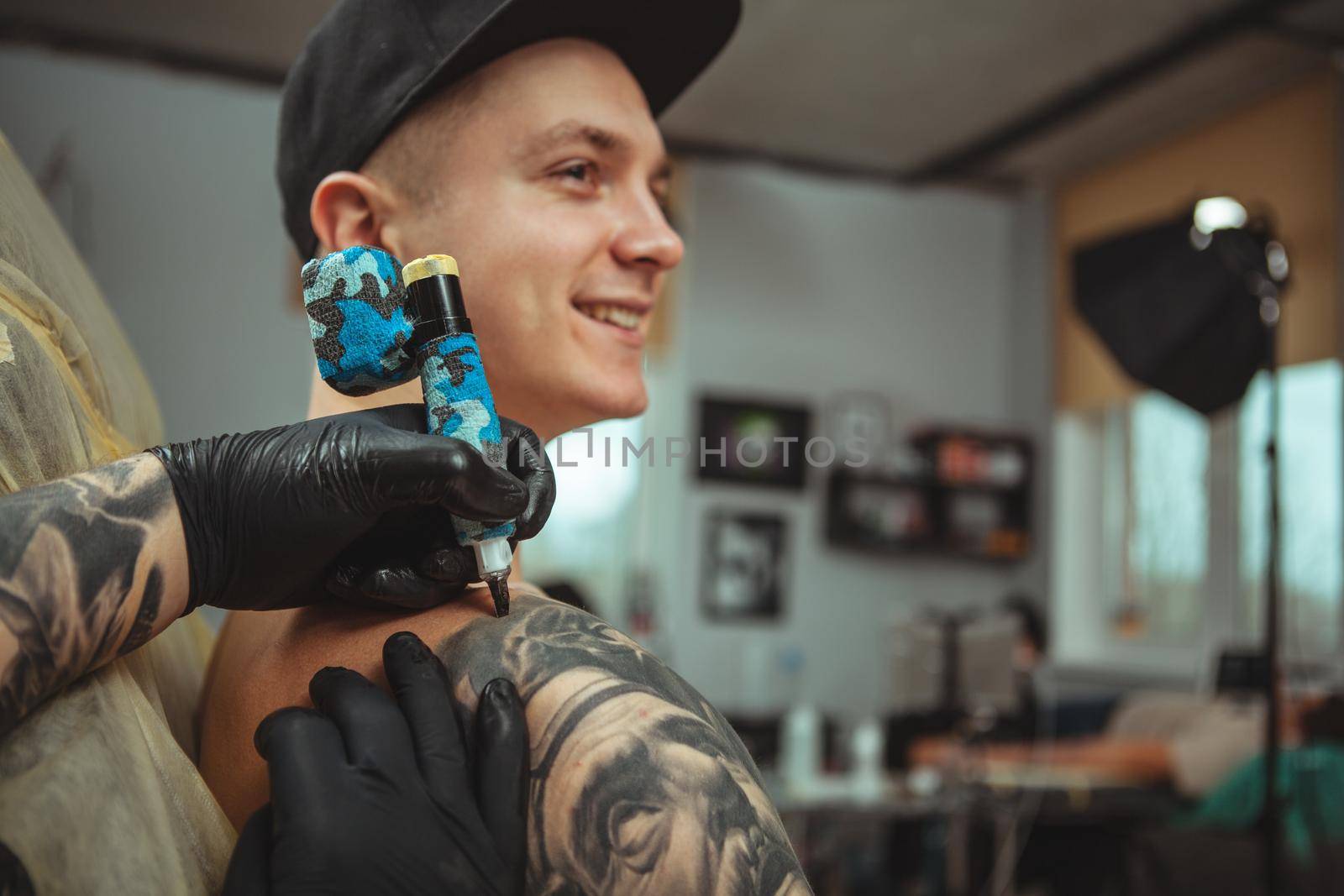 Cropped shot of hands of professional tattoo artist making a tattoo on the shoulder of his male client. Young man smiling, getting tattooed by professional tattooist