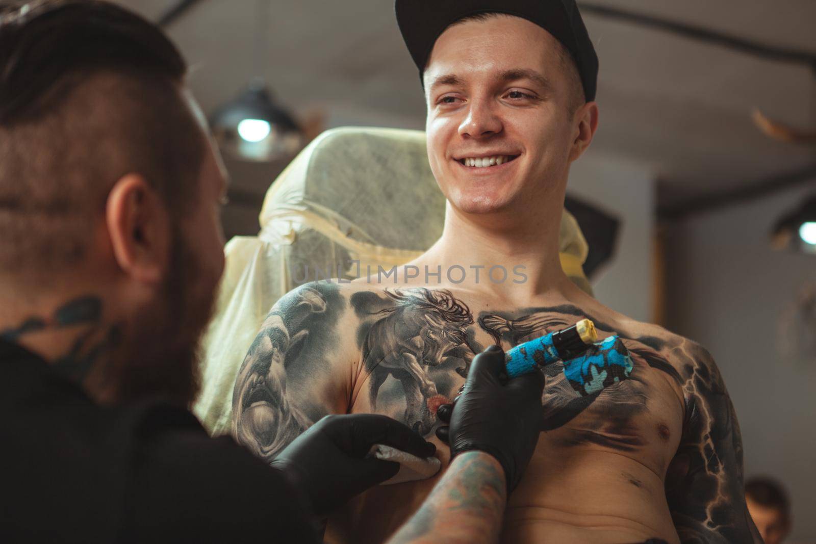 Handsome cheerful young man smiling, talking to his tattoo artist while getting inked at salon. Attractive male client getting chest tattoo by professional tattooist. Service, consumerism concept