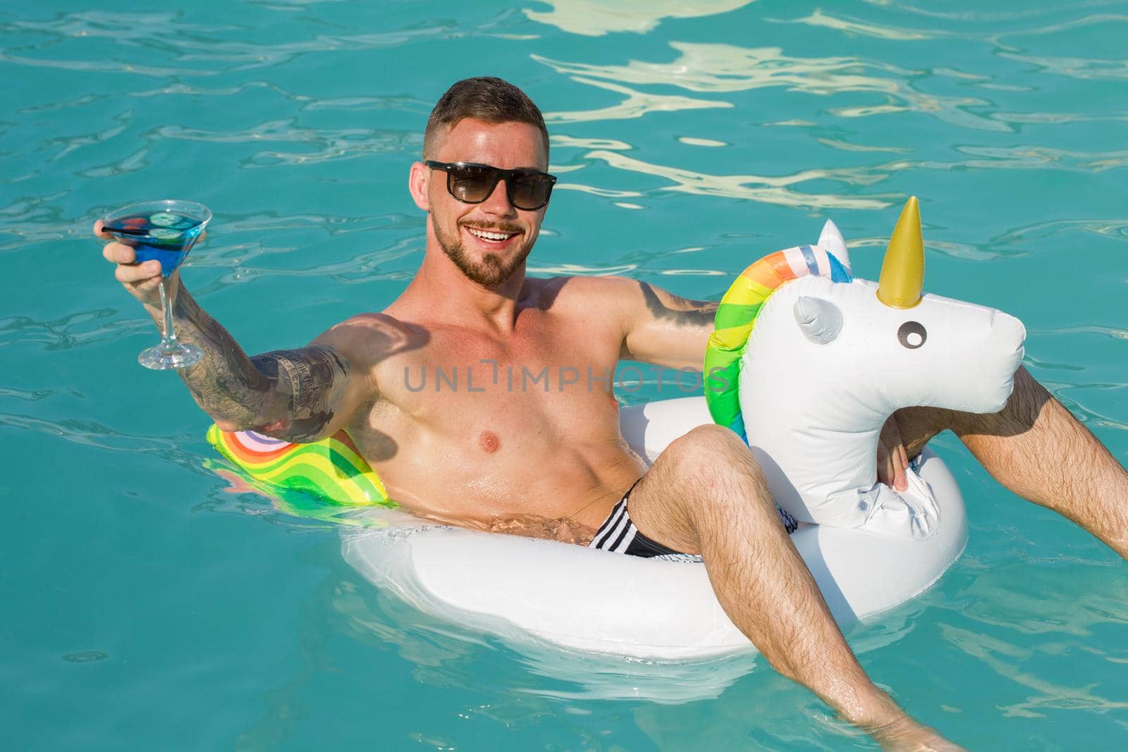 Cheerful handsome bearded man cheering with his cocktail glass while swimming on inflatable unicorn at the swimming pool. Happy man relaxing during his summer vacation. Lifestyle, partying, travel concept