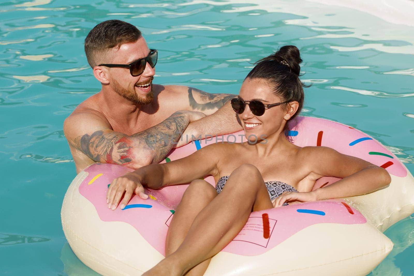 Happy couple talking while swimming on inflatable donut at the pool. Beautiful woman sitting on pool float, chatting with her boyfriend, relaxing together during summer vacation. Love, travel, lifestyle concept