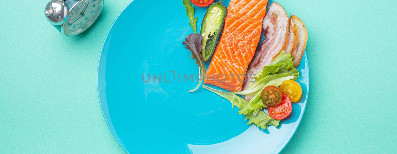 Intermittent fasting low carb hight fats diet concept flat lay, healthy food salmon fish, bacon meat, vegetables and salad on blue plate and clock alarm on blue background top view