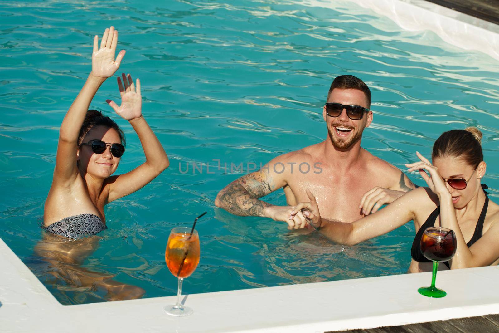 Group of friends dancing in the water at the pool party. Cheerful young people having drinks, laughing joyfully, enjoying hot summer day at the swimming pool. Entertainment, happiness, travel concept
