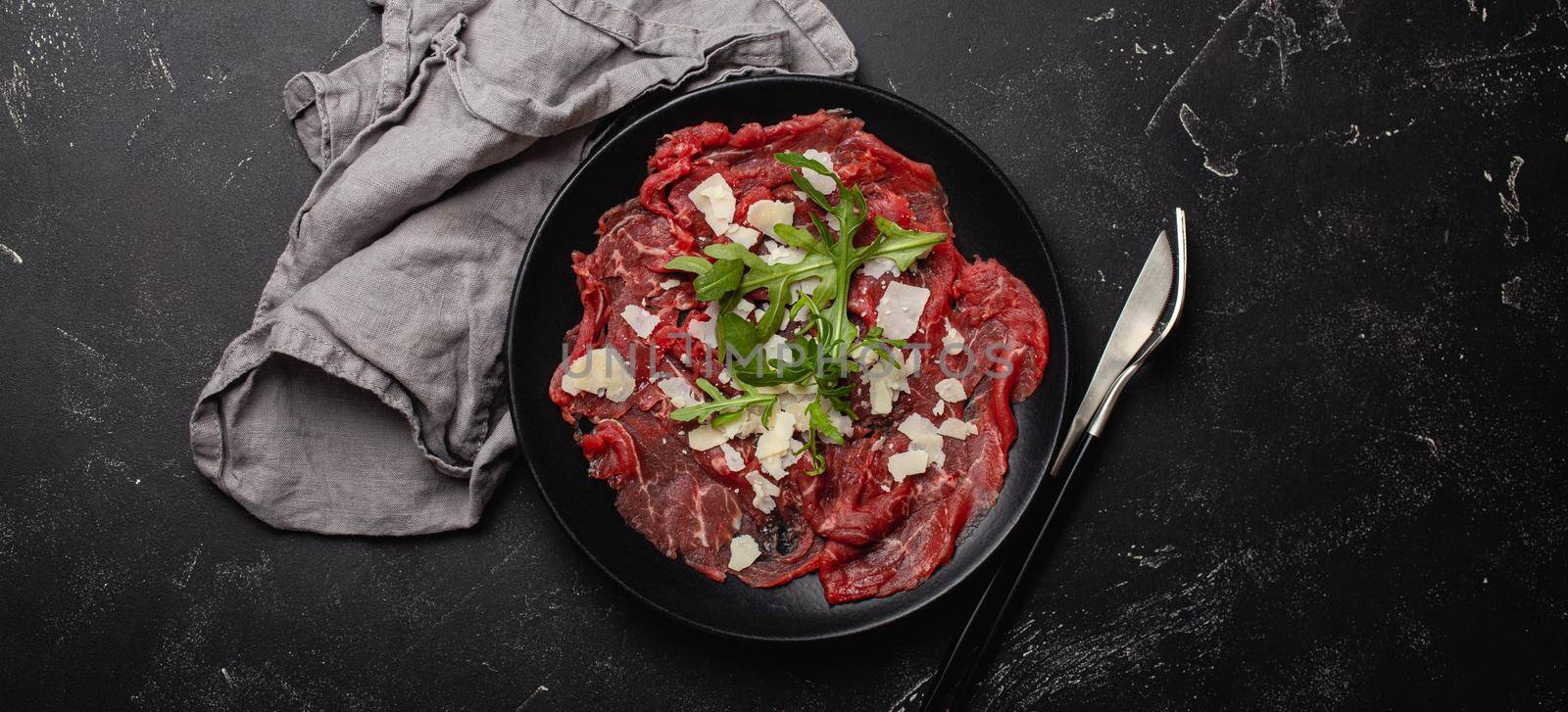 Italian cold meat appetizer Beef carpaccio with parmesan cheese and arugula on black plate with fork and knife on dark stone concrete rustic background flat lay from above .