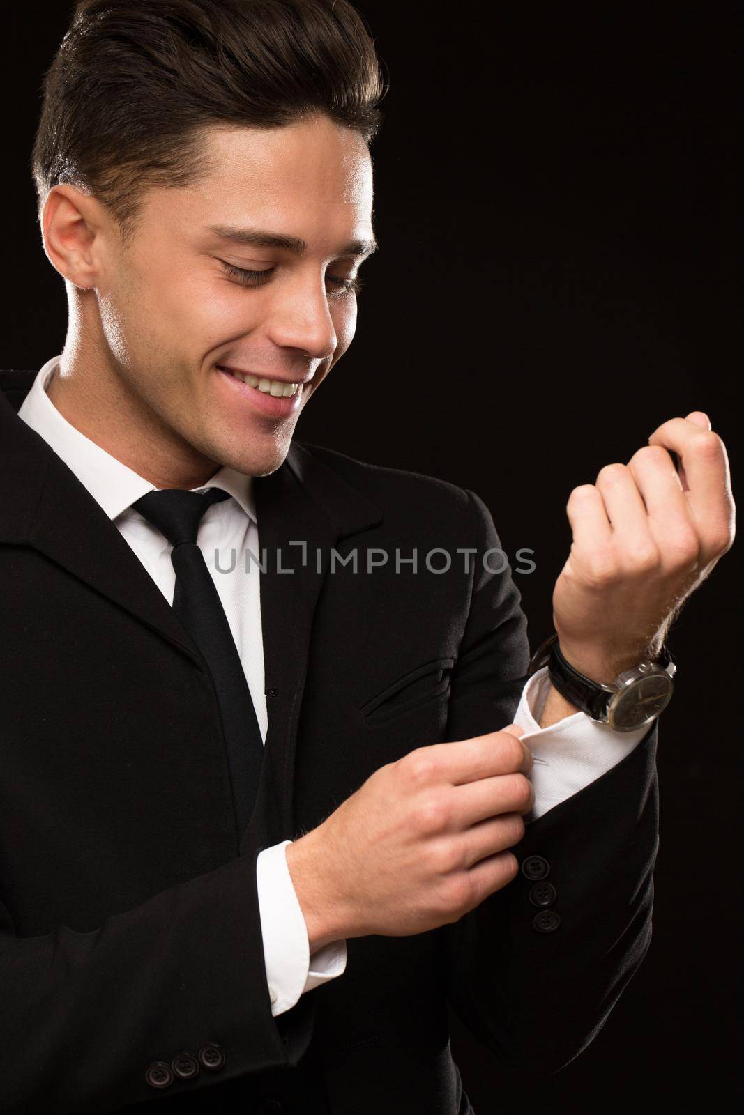 Vertical cropped shot of a handsome young suited man smiling joyfully adjusting sleeves of his shirt on black background macho positivity confidence success achievement businesspeople style