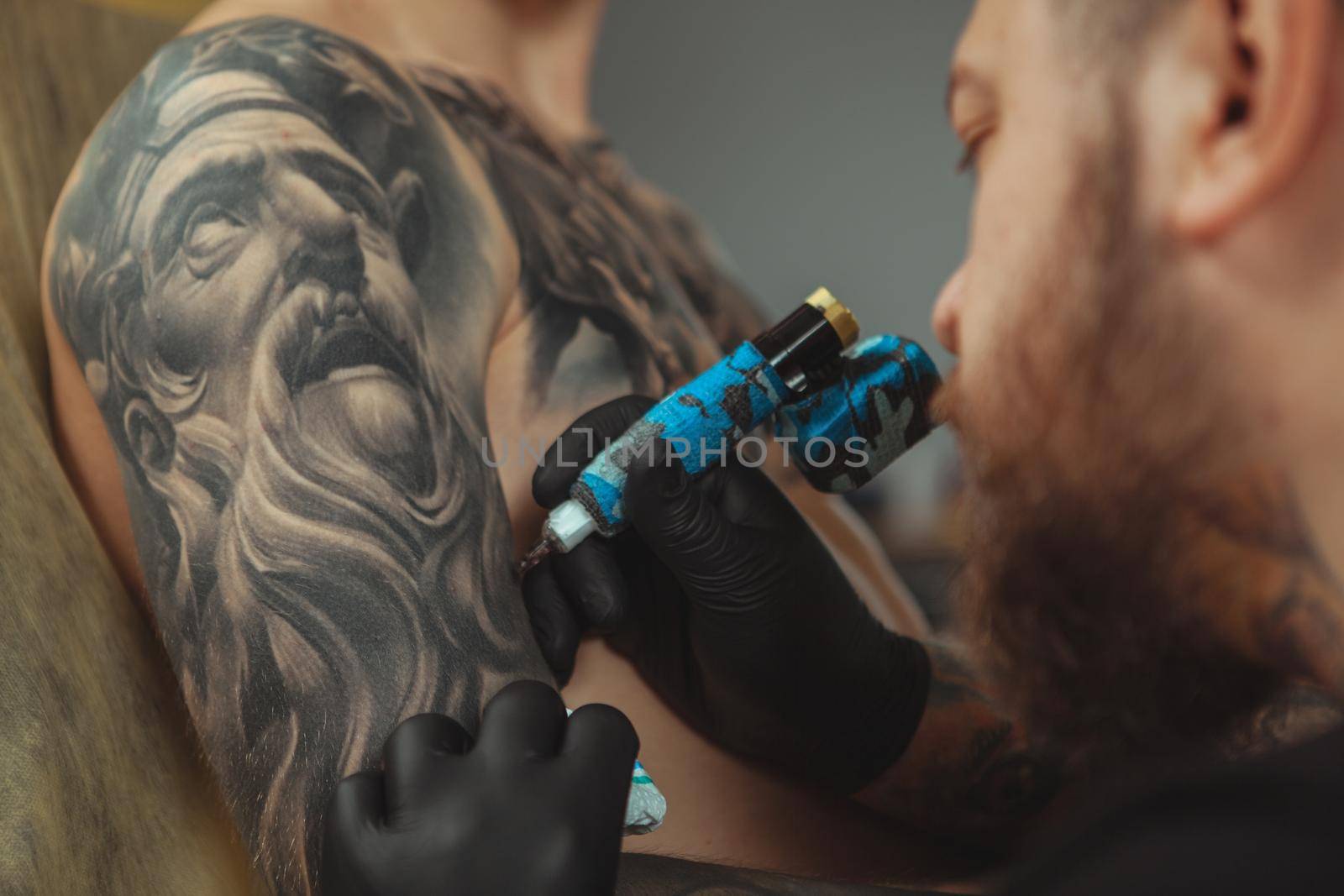 Cropped close up of a bearded tattoo artist working at his studio tattooing sleeve on the arm of his male client. Man getting tattooed by professional tattooist