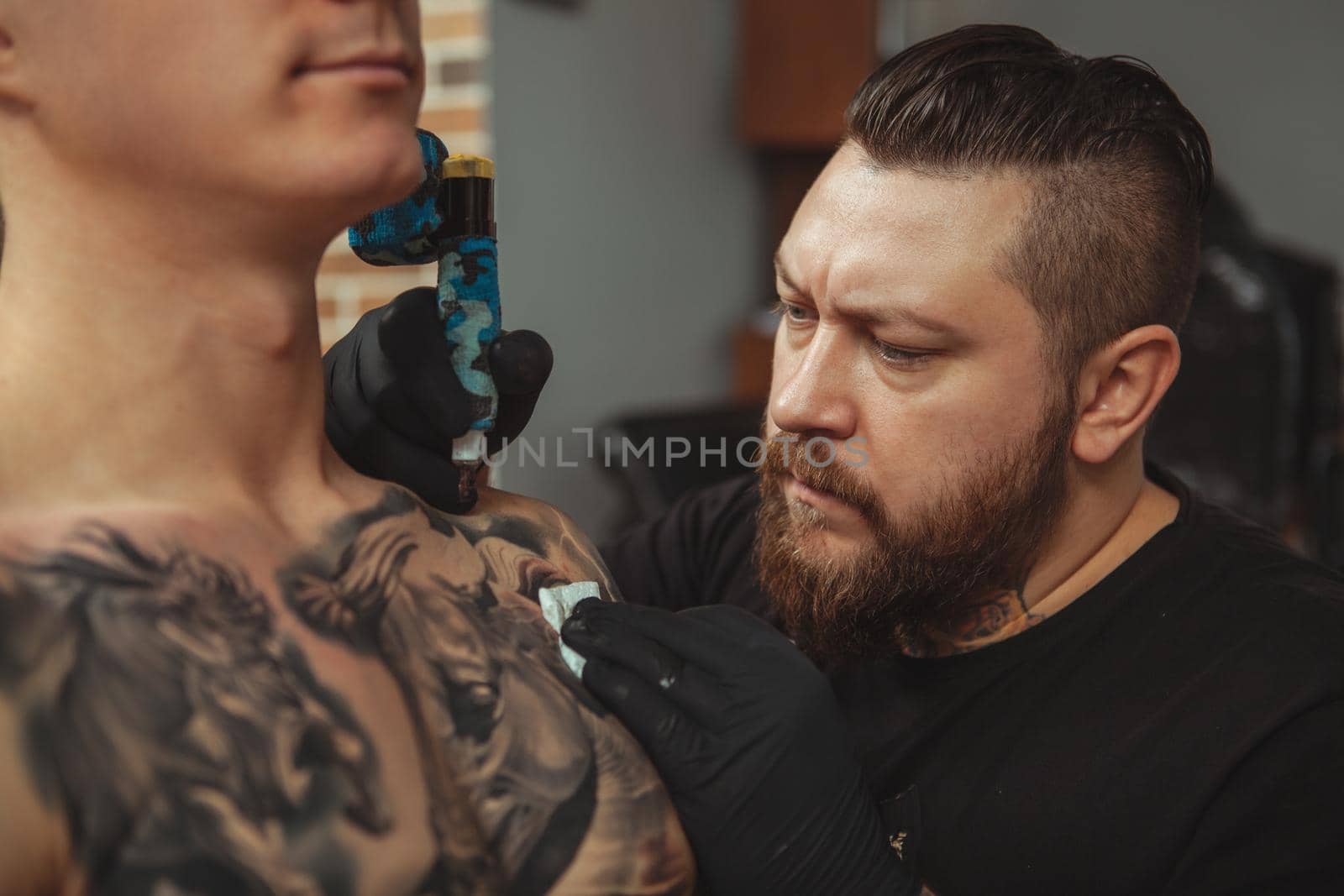 Close up of a concentrated bearded tattoo artist working, tattooing shoulder of a male client. Professional tattooist looking focused, copy space