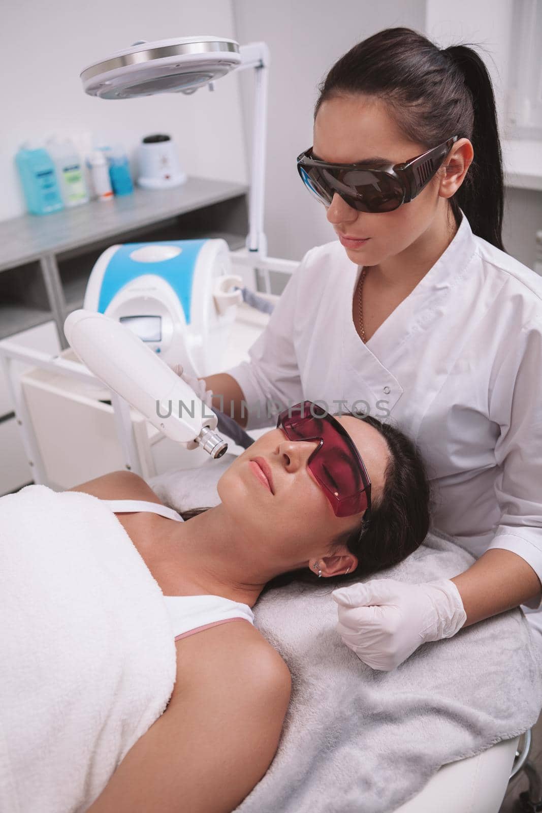 Vertical shot of a professional beautician and female client wearing protective glasses during facial hair laser removal procedure