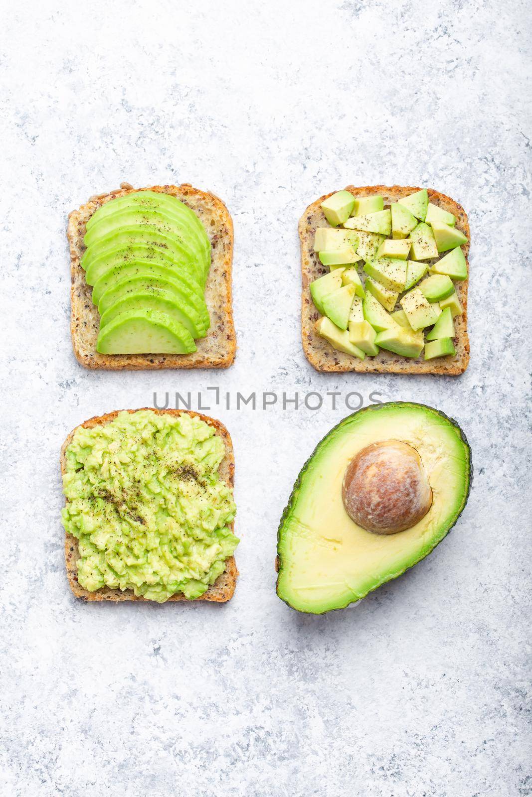 Different types of avocado toasts and a half of whole avocado over white stone background, concept. Ways of making and serving delicious healthy breakfast avocado sandwich, top view, close-up.