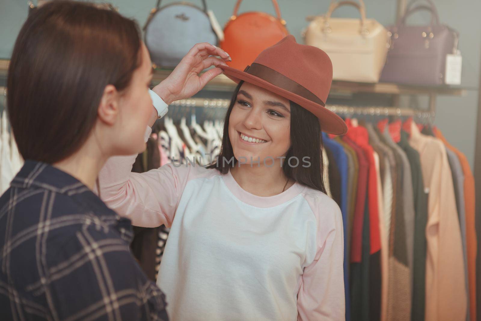 Beautiful cheerful young woman trying on a hat at clothing store, talking to her friend, copy space. Two young women enjoying shopping for clothing and accessories together