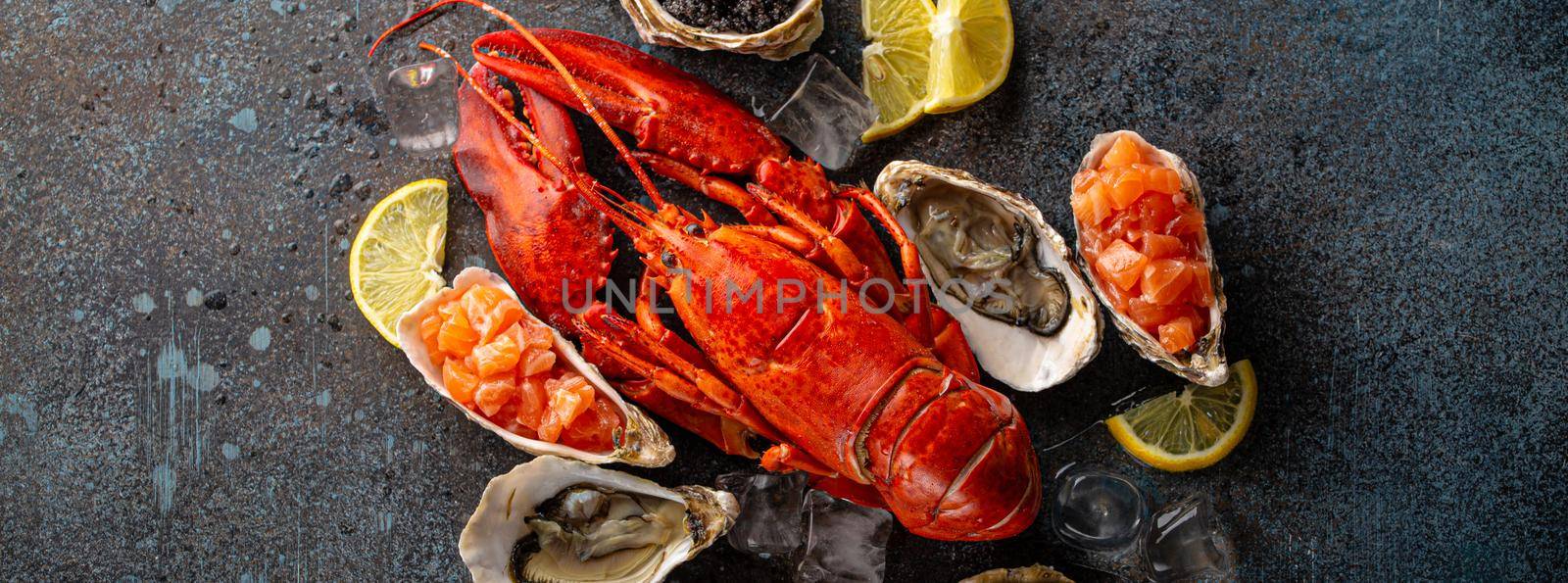 Seafood assorted plate with cooked red whole lobster, fresh open oysters, black caviar, salmon tartare served with lemon wedges and ice cubes top view flat lay on blue concrete stone background