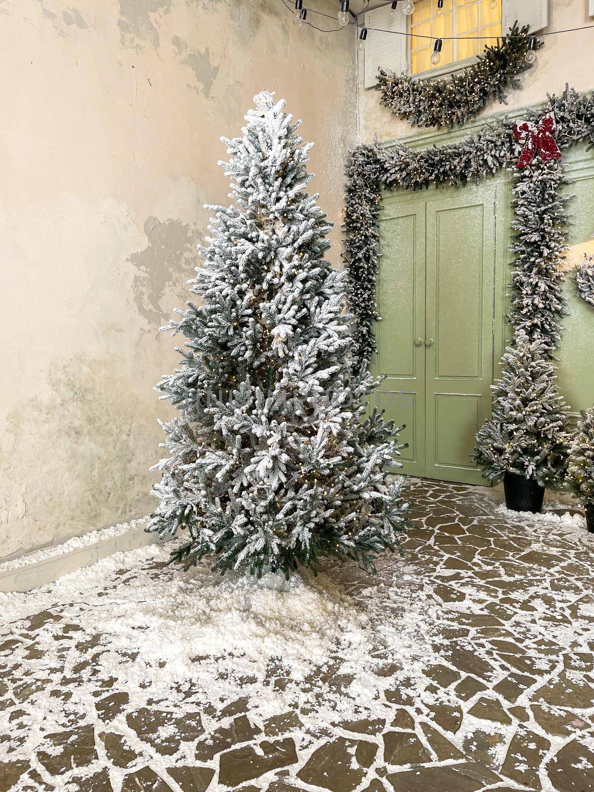 Christmas tree covered in snow and decorated with garland outside next to front door of cafe in pastel green tones with wreaths, lanterns, windows and many fir trees in snow