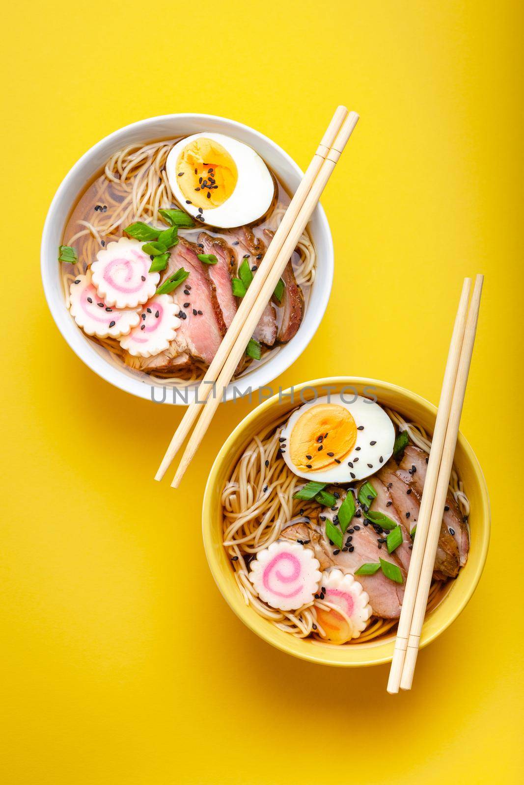 Two bowls of Japanese noodle soup ramen with meat broth, sliced pork, narutomaki, egg with yolk on pastel yellow background. Traditional dish of Japan, top view, close-up, concept