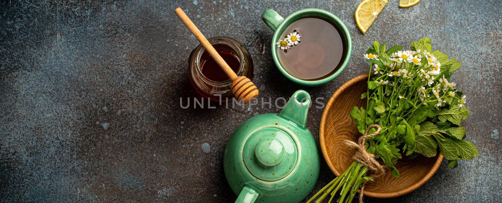 Herbal tea with camomile, fresh medical herbs and flowers, tea cup, tea pot, jar of honey, lemon top view on stone background, herbal alternative medicine and homeopathy remedy concept space for text