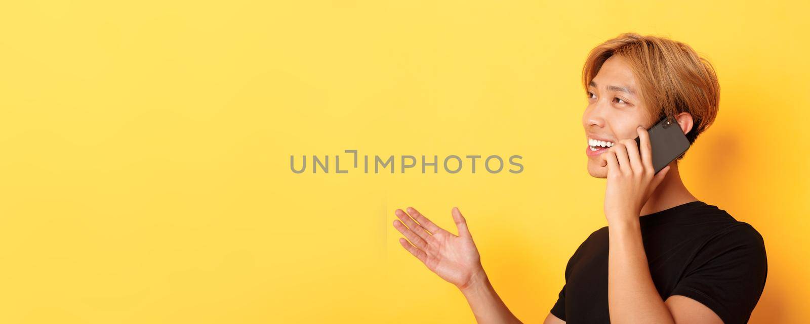 Close-up of attractive korean guy smiling and talking on mobile phone happy, turning left, standing yellow background by Benzoix