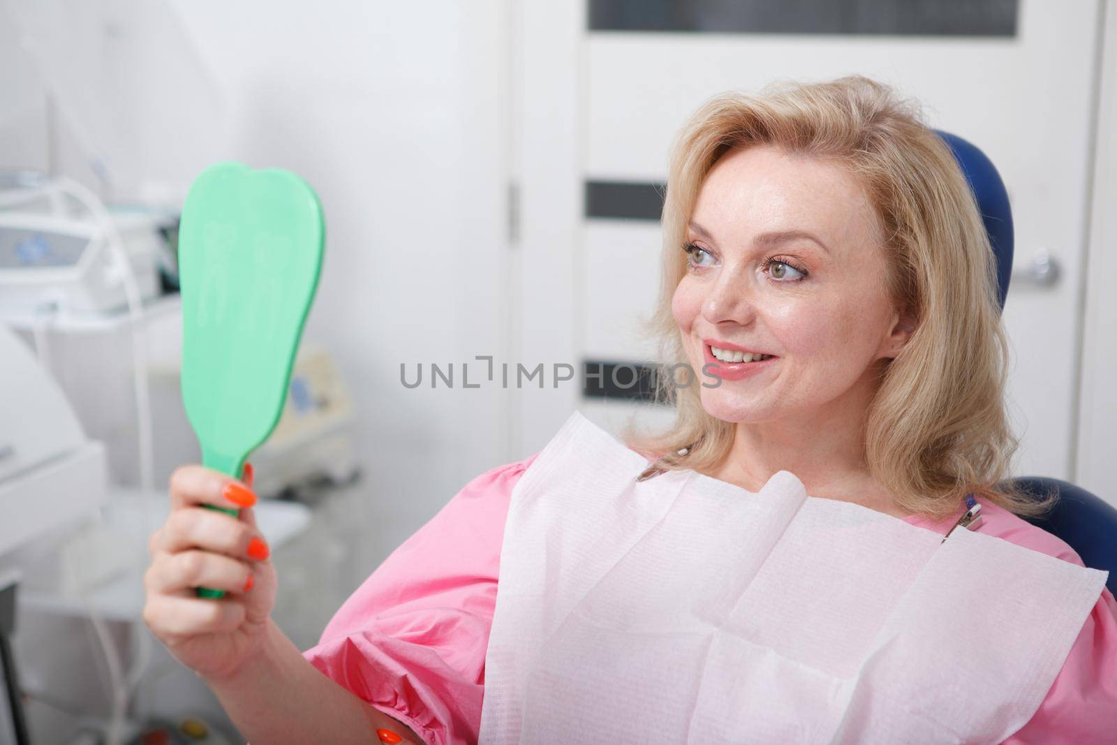 Cheerful mature woman smiling to the mirror she is holding, checking teeth after dental treatment