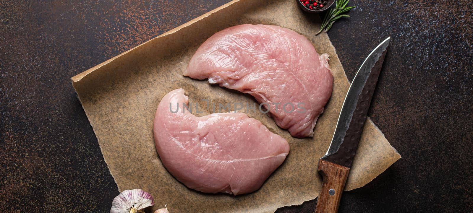 Turkey lean raw fillet on baking paper with rosemary, garlic and spices on dark brown rustic concrete background with knife from above flat lay, healthy diet turkey meat ready to be cooked