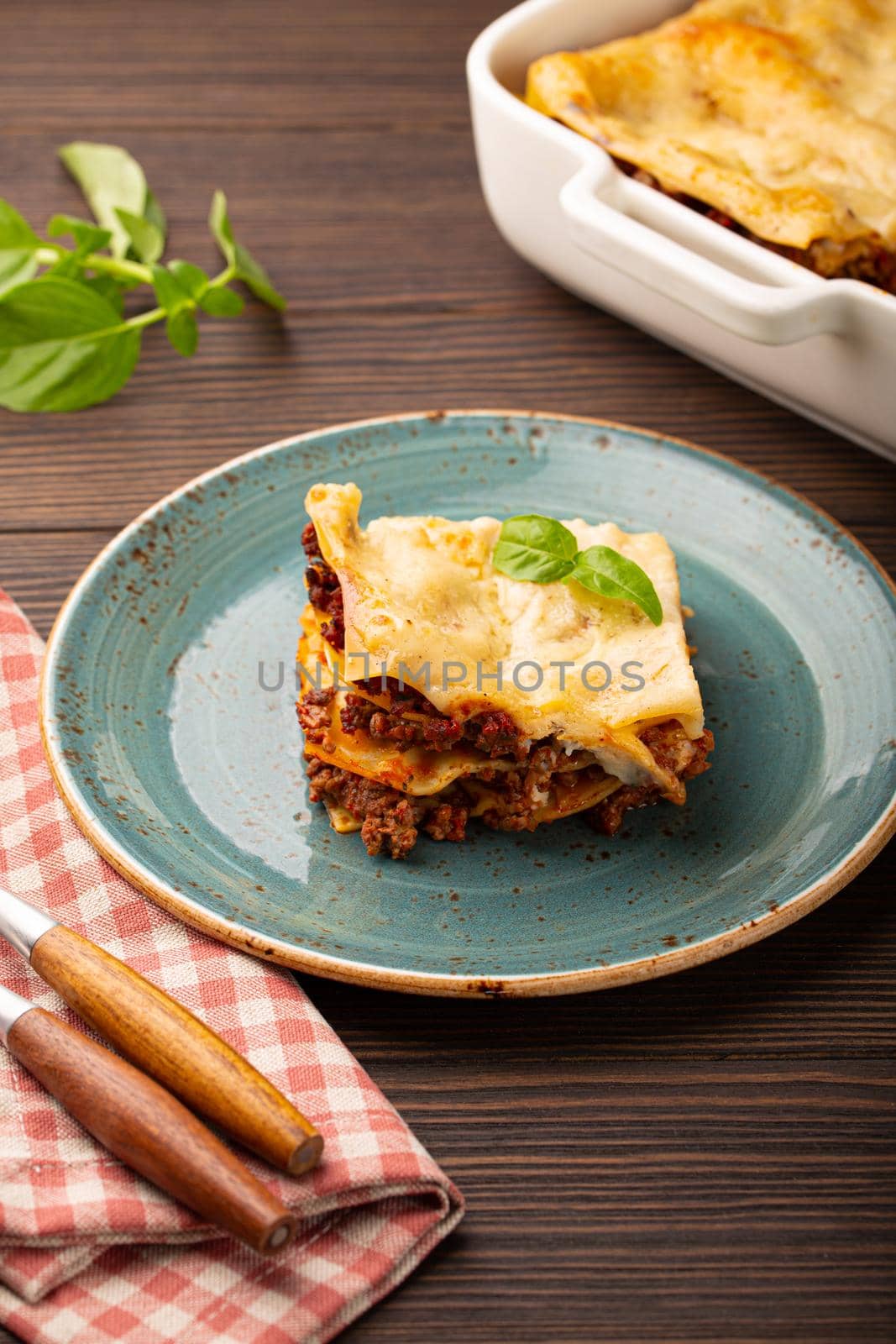 Slice of homemade traditional Italian lasagna with beef bolognese ragout and cheese served on blue ceramic plate on dark brown rustic wooden table, angle view