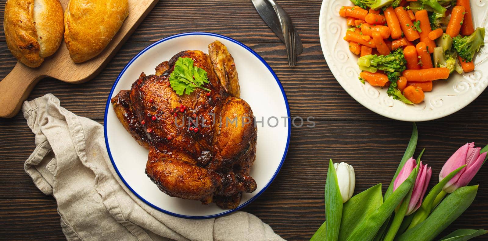 Easter food on dark rustic table: pastel colored eggs, roasted chicken and vegetables, buns and spring flowers tulips top view flay lay, Easter family dinner meal with festive dishes concept