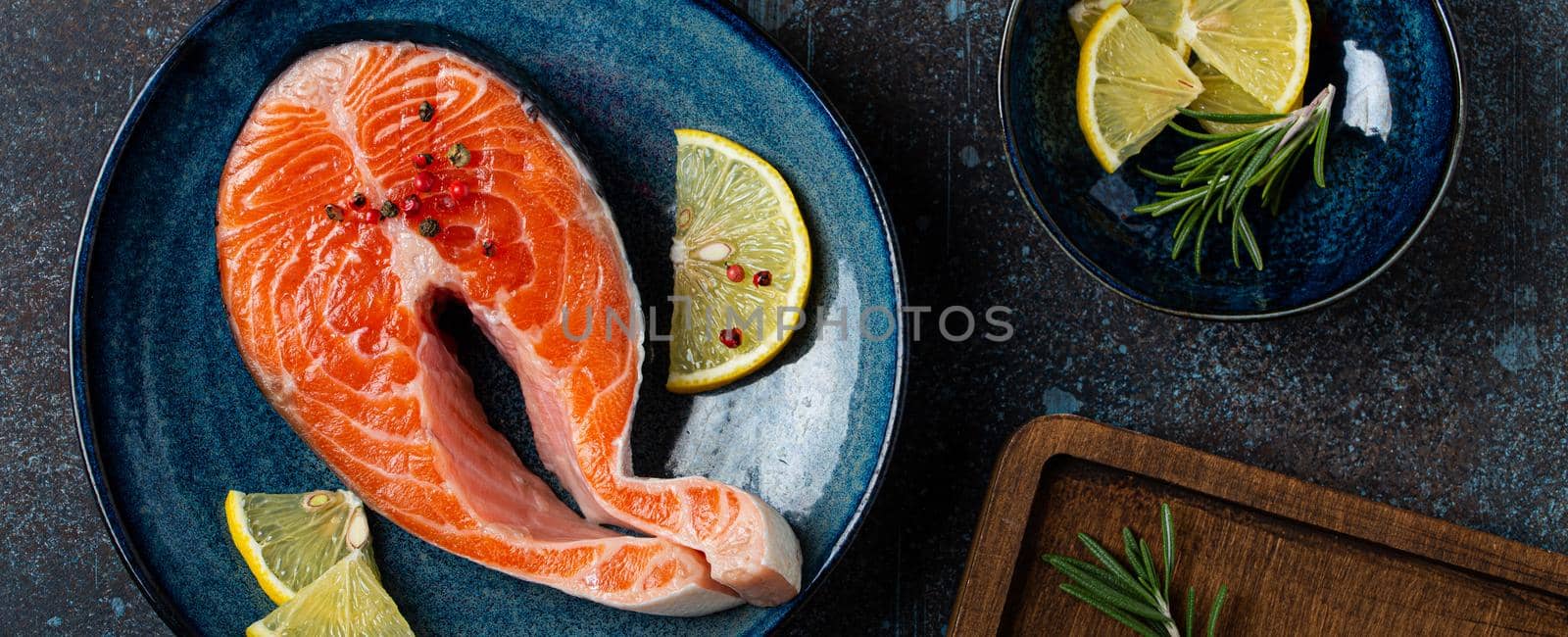 Uncooked raw fresh fish salmon steak top view on plate rustic dark concrete stone background with rosemary, lemon wedges and spices, delicacy healthy fish cooking and nutrition concept flat lay