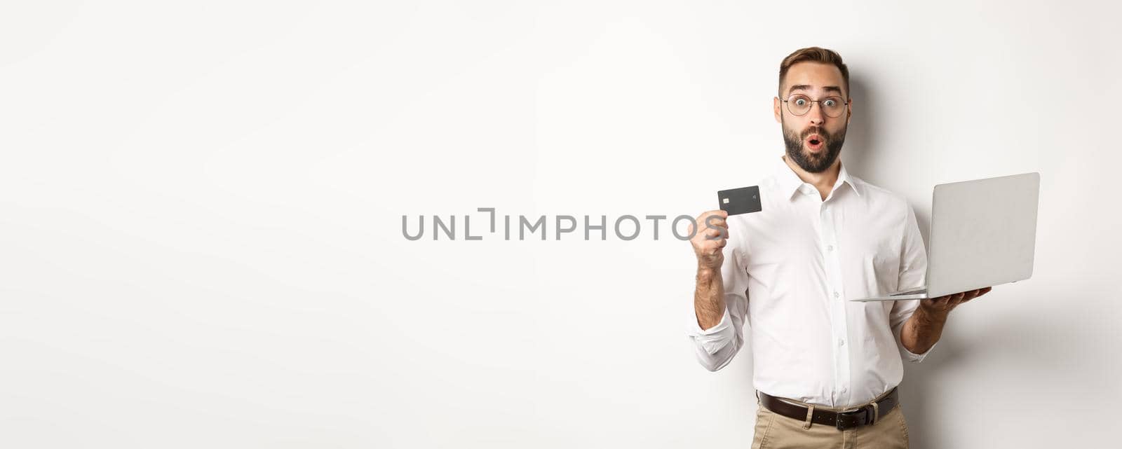 Online shopping. Surprised man holding laptop and credit card, shop internet store, standing over white background.