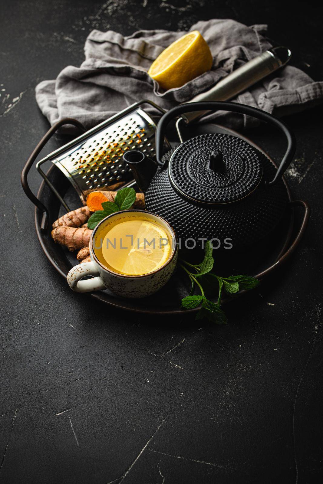 Healthy warm herbal turmeric tea for boosting immune system. Ingredients for detox tea in cup, black teapot, fresh curcuma root, lemon, mint leaves, grater on black background, top view, copy space.