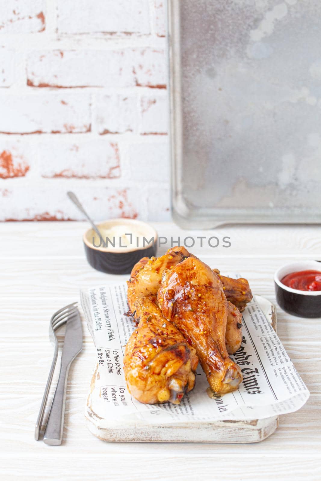 Roasted chicken legs drumsticks on wooden board with newspaper sheet served with ketchup and mayonnaise on rustic white wooden table, angle view
