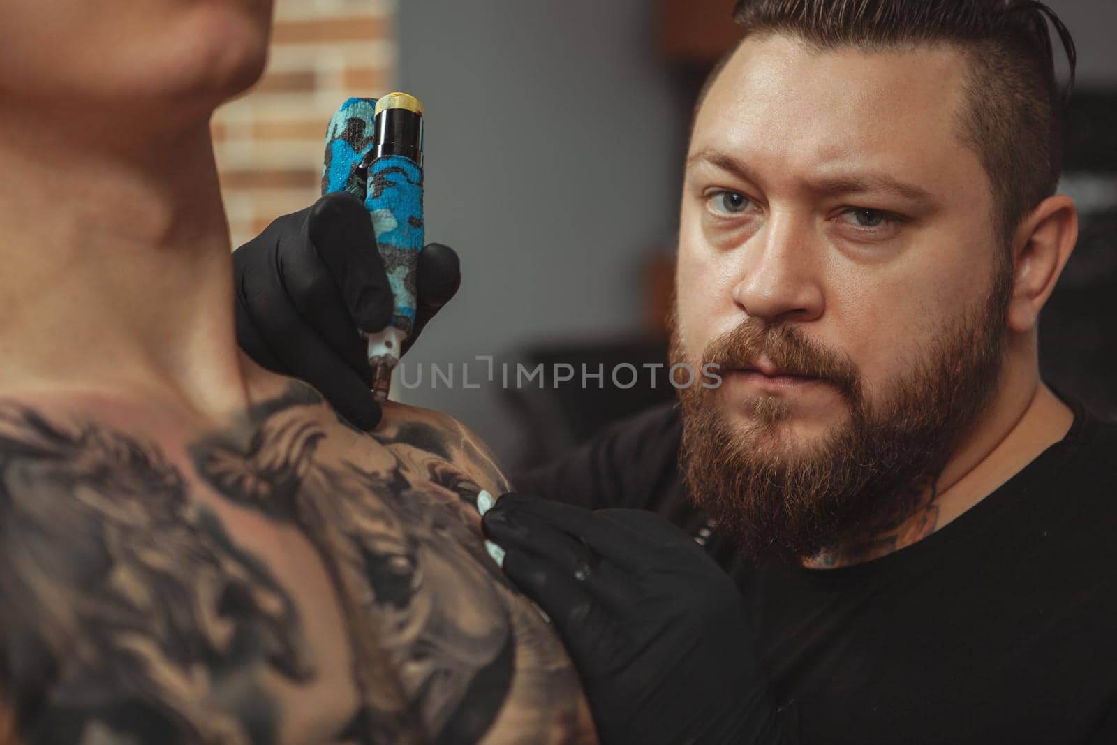 Cropped close up of a professional tattoo artist looking to the camera fiercely while working. Experienced tattooist tattooing chest of a male client. Professionalism, creativity concept