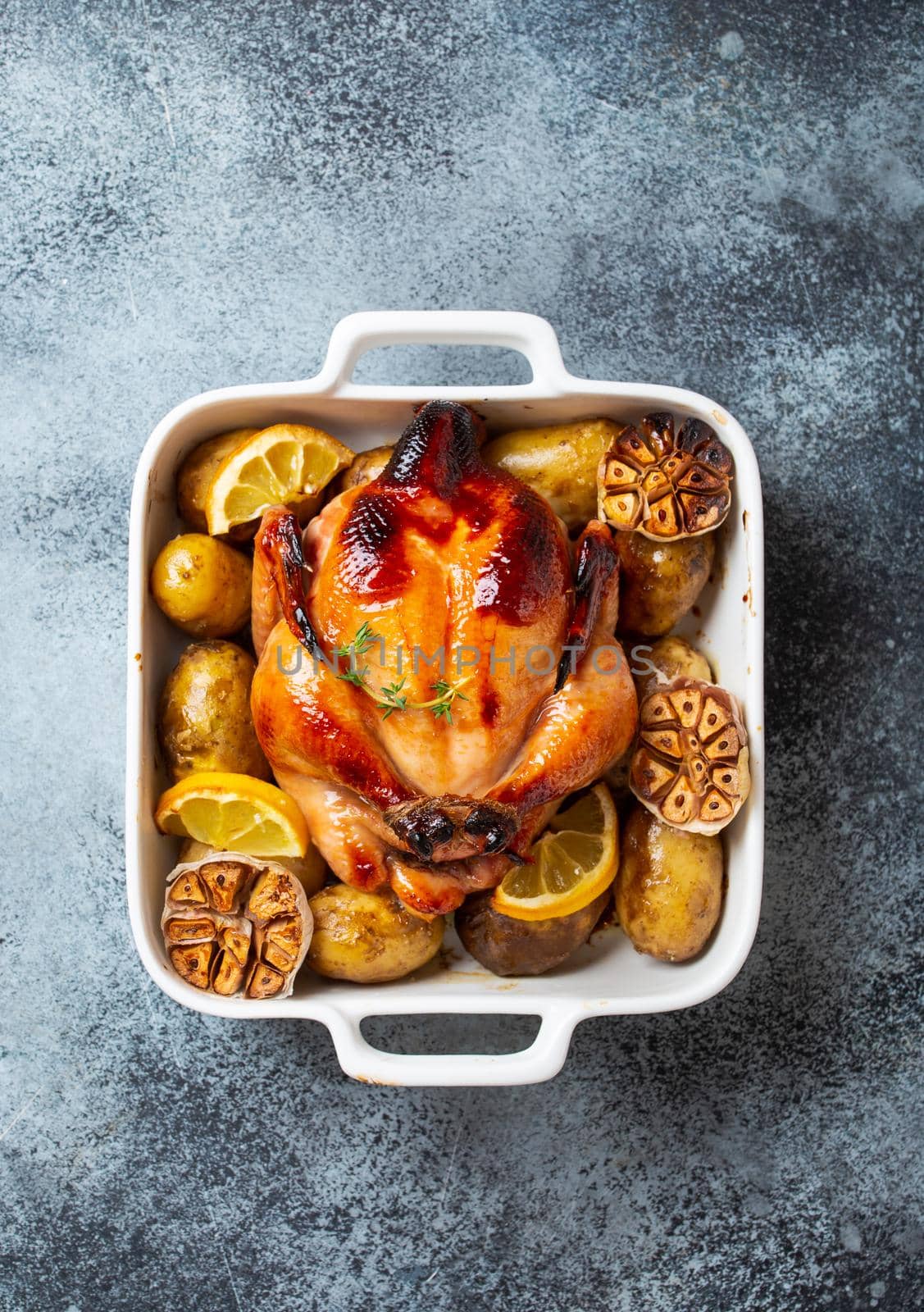 Close-up of baked or roasted whole juicy crispy organic chicken with potatoes, garlic, lemon in white casserole dish on concrete rustic background, top view