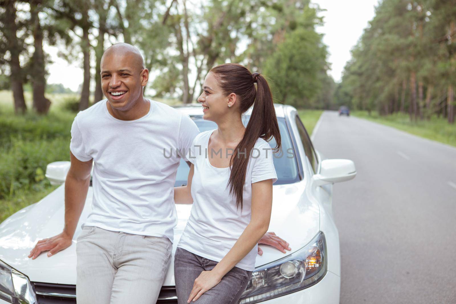 Charming multiracial couple enjoying travelling together by car. Handsome African man hugging his beautiful girlfriend, leaning on a car on the side of the road