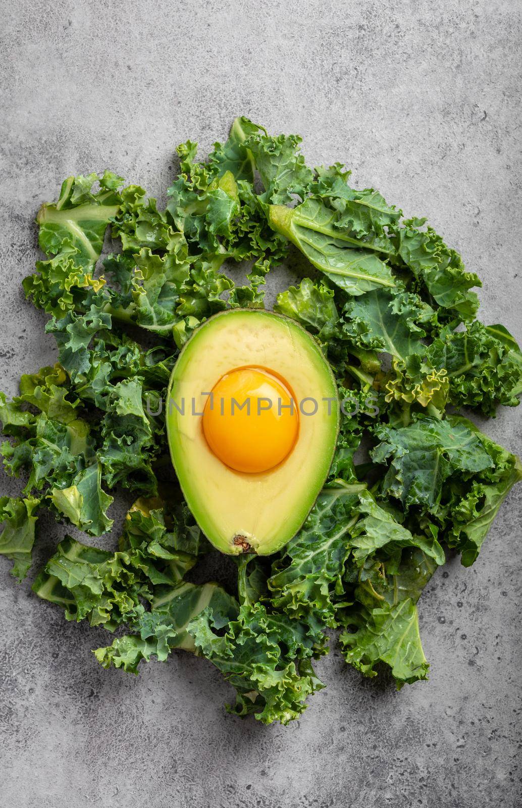 Raw egg yolk in fresh cut half avocado rich in healthy fats and kale leaves for balanced menu on gray stone background. Ketogenic low carbs diet or clean eating concept, top view .