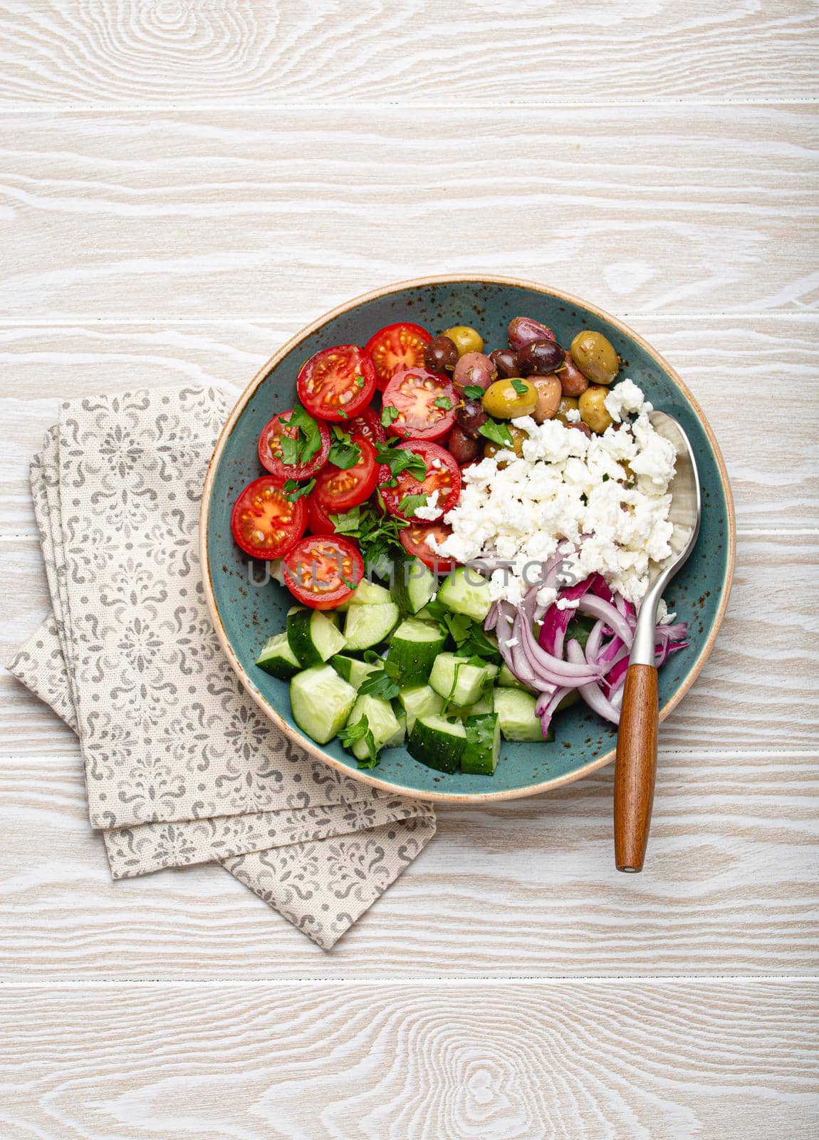 Greek mediterranean salad with tomatoes, feta cheese, cucumber, whole olives and red onion in blue ceramic plate on white wooden background from above, traditional appetizer of Greece