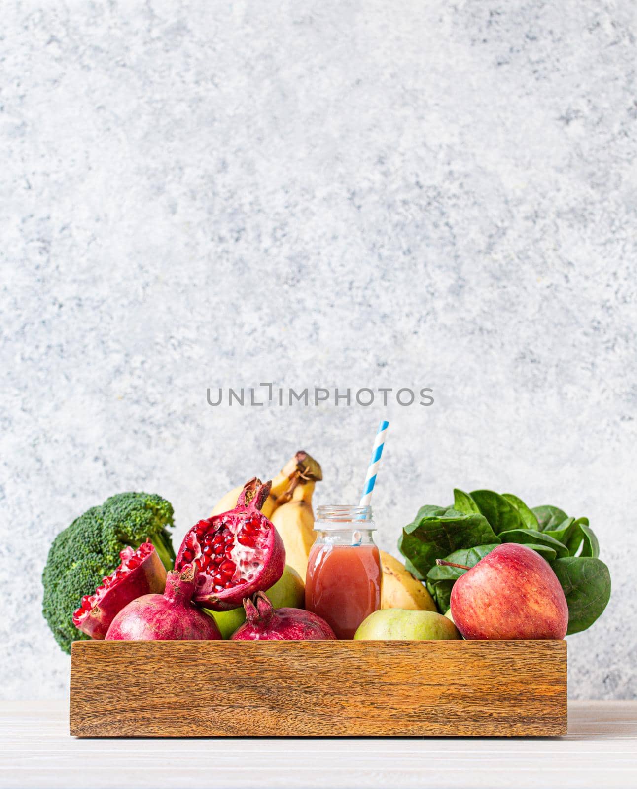 Fruit, greens, vegetables in wooden tray and fresh smoothie or juice in bottle with paper straw on table near white wall, detox, diet, clean eating, vegetarian, healthy food concept, space for text.