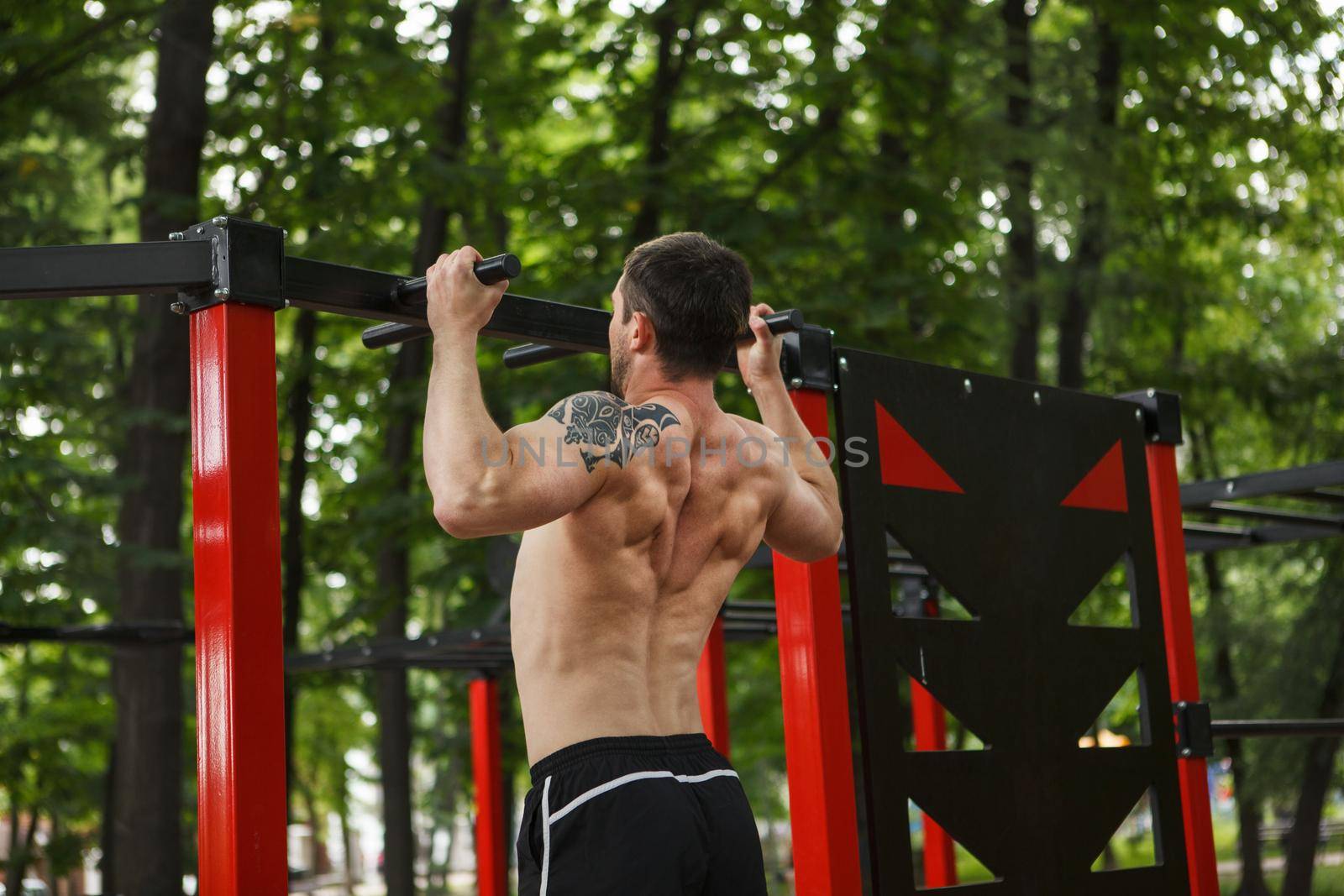 Shirtless muscular male athlete with ripped torso doing ull ups outdoors