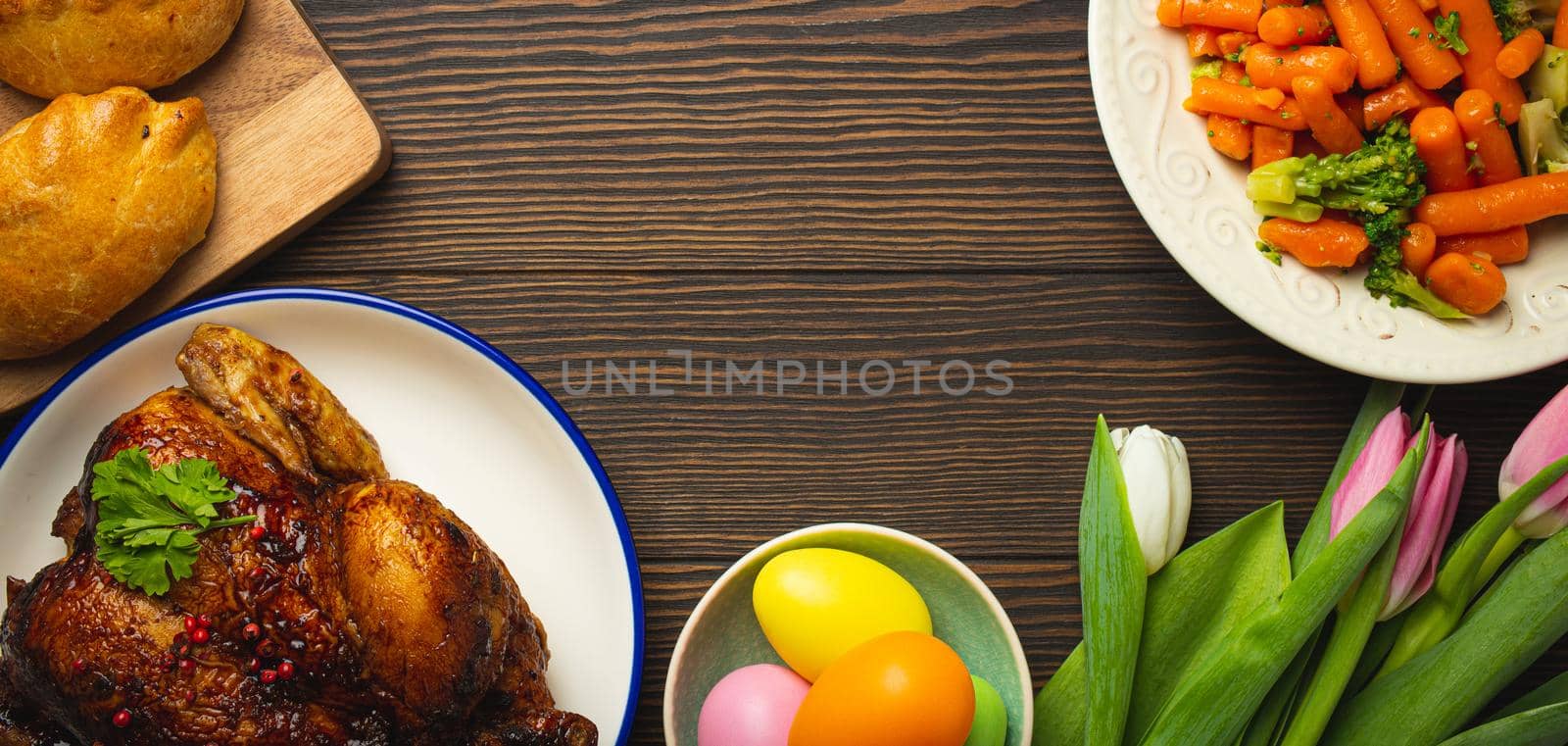 Easter food on dark rustic table: pastel colored eggs, roasted chicken and vegetables, buns and spring flowers tulips top view flay lay, Easter family dinner meal with festive dishes, space for text
