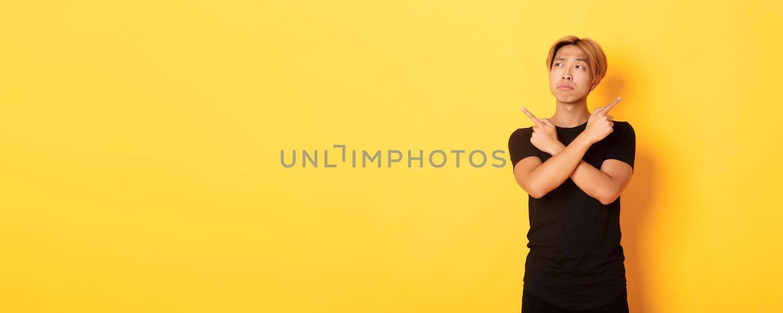 Indecisive handsome asian guy looking confused, pointing fingers sideways, standing yellow background.