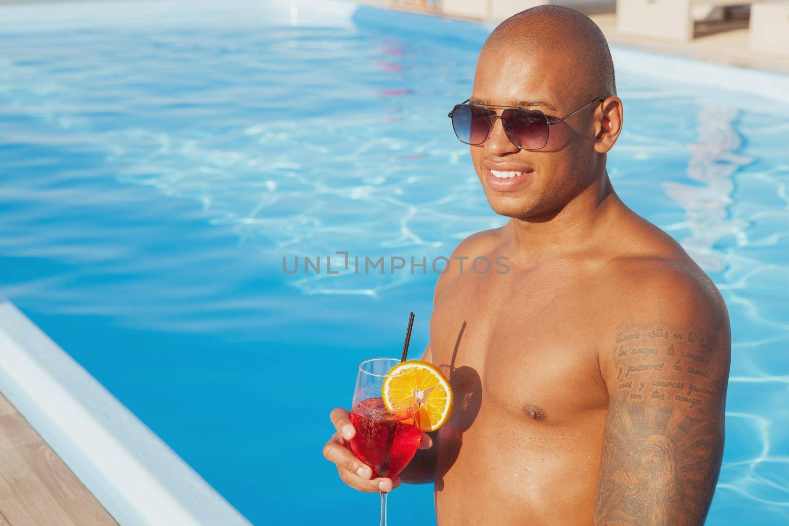 Handsome happy African man smiling, looking away thoughtfully, relaxing at poolside. Attractive man enjoying cocktail at the swimming pool