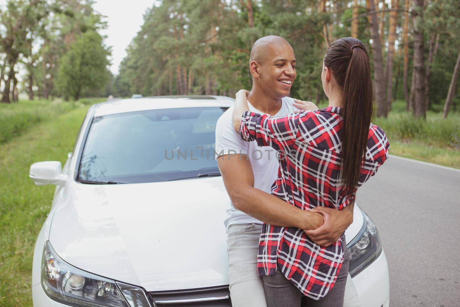 Young happy couple hugging near their car, taking a break from driving during roadtrip, copy space. Lovely multiracial couple cuddling on the side of the road near auto